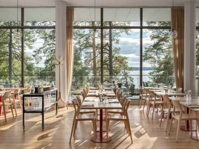 Bilden förställer Artipelag Restaurang i solljus. På bilden syns dukade bord på rad framför ett panoramafönster med utsikt över vattnet och talltopparna.