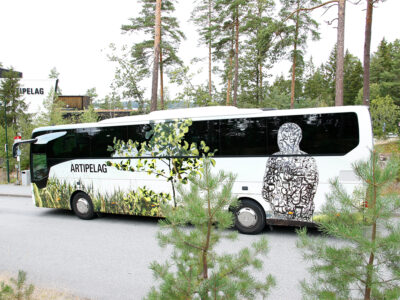 Bild på en buss folierad med Artipelagmotiv så som en skulptur av konstnären Jaume Plensa. Bussen står parkerad på Artipelag, i förgrunden skymtar några grankvistar och i bakgrunden ser man tallar och en del av byggnaden.