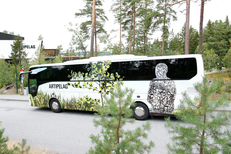 Bild på en buss folierad med Artipelagmotiv så som en skulptur av konstnären Jaume Plensa. Bussen står parkerad på Artipelag, i förgrunden skymtar några grankvistar och i bakgrunden ser man tallar och en del av byggnaden.