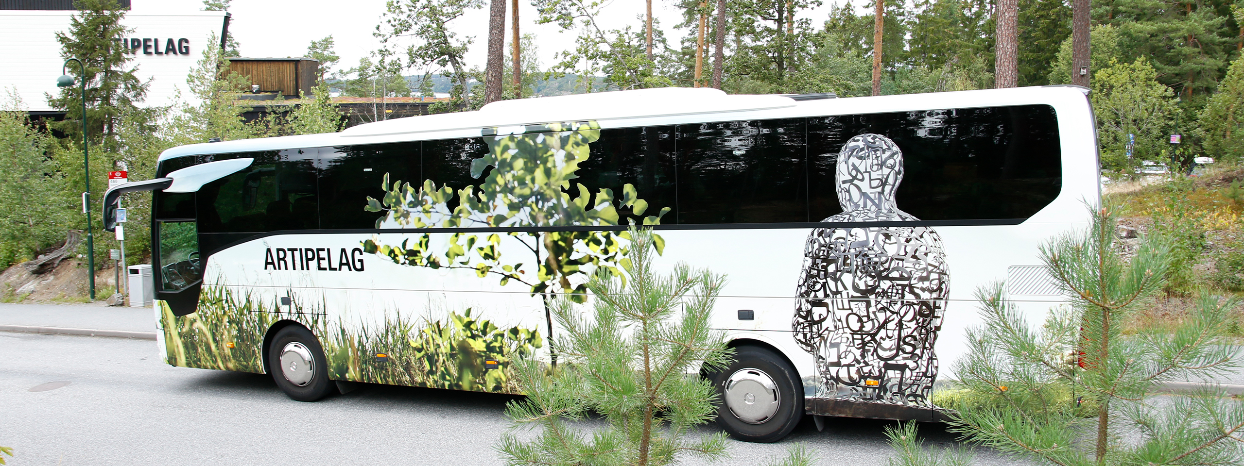 En buss av märket "ARTIPELAG" visar upp konstnärliga mönster av grönska och en figur gjord av sammanlänkade former, parkerad på en lugn väg omgiven av träd.