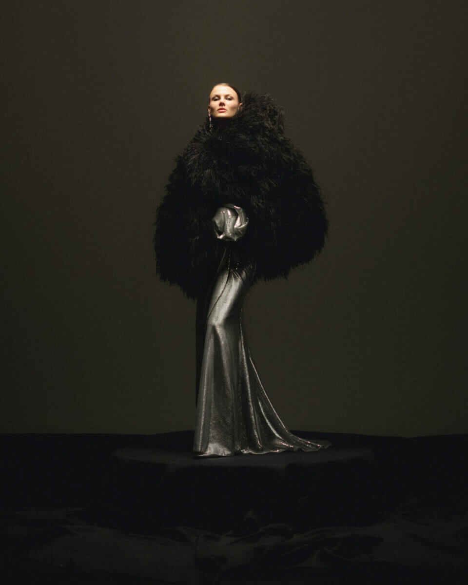 En person är elegant utsmyckad i en dramatisk, stor svart fjäderkappa över en metallisk silverklänning, mot en mörk bakgrund. Denna fantastiska ensemble fångar essensen av Lars Wallin Atelier – 35 Years of Fashion Stories