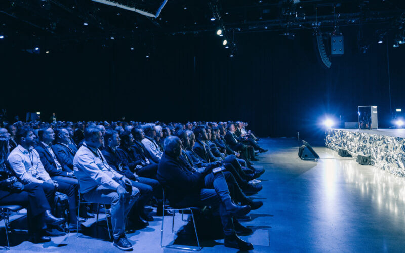 Publiken sitter i en svagt upplyst auditorium, vänd mot en scen med starkt ljus.