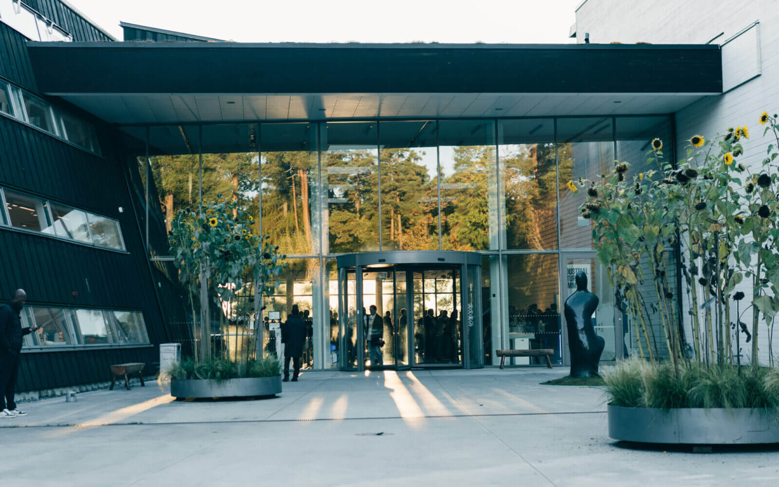 Modern glasbyggnadsingång med svängdörr, flankerad av geometriska väggar och höga krukväxter. Människor är synliga genom glaset och solljus kastar långa skuggor på marken.