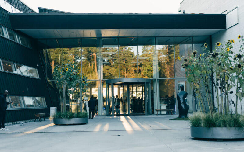 Modern glasbyggnadsingång med svängdörr, flankerad av geometriska väggar och höga krukväxter. Människor är synliga genom glaset och solljus kastar långa skuggor på marken.