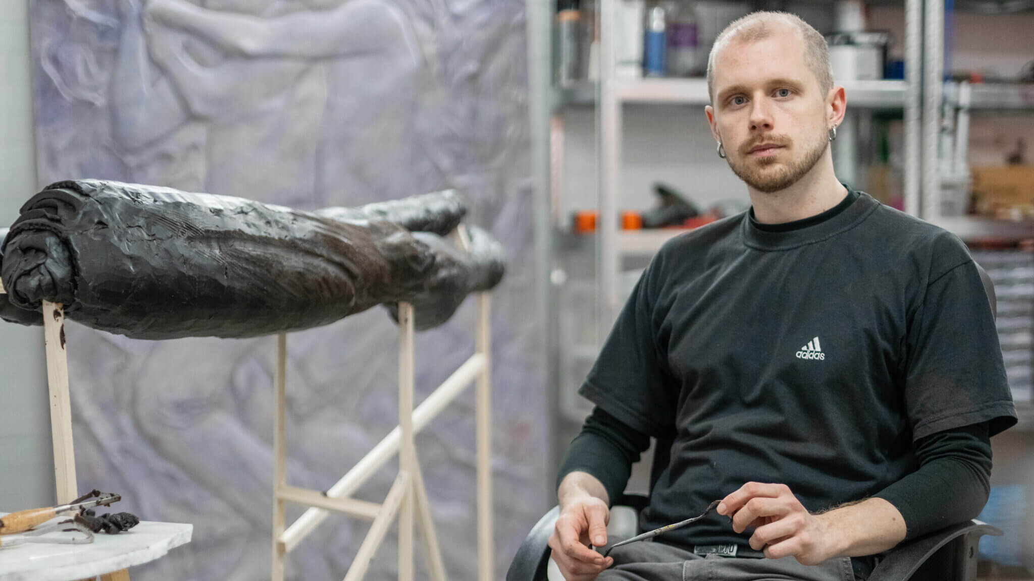 En man i svart t-shirt sitter bredvid ett stort, mörkt skulpturalt konstverk i en ateljé med konstnärsmaterial och en målning i bakgrunden, möjligen under förberedelse inför Fredrik Roos stipendium 2025.