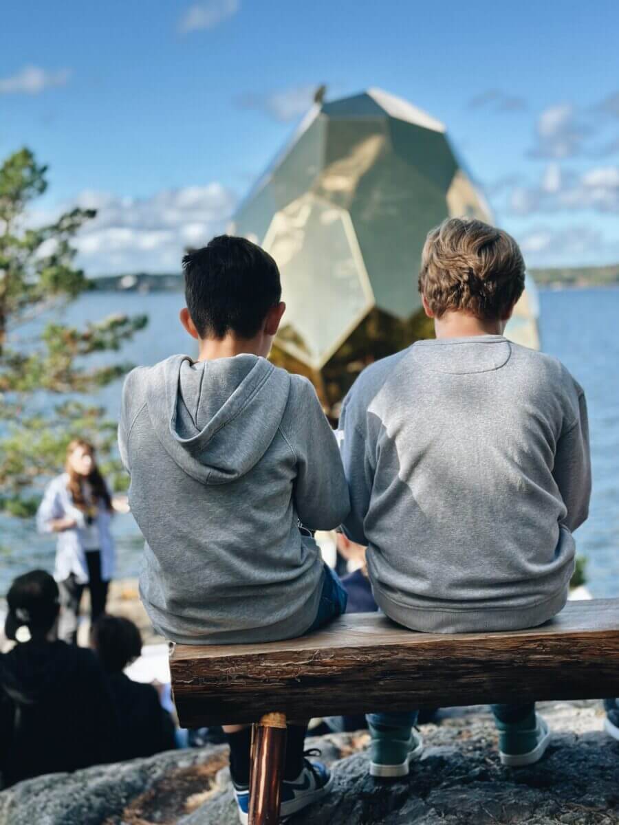 Två barn som sitter på en bänk framför havet och framför en stor guldfärgad och äggformad skulptur.